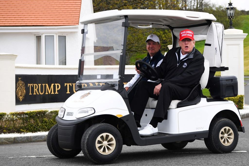 Trump, wearing a windbreaker and a red MAGA hat, speaks from the driver's seat of a golf cart. A TRUMP TURNBERRY sign is visible behind him.