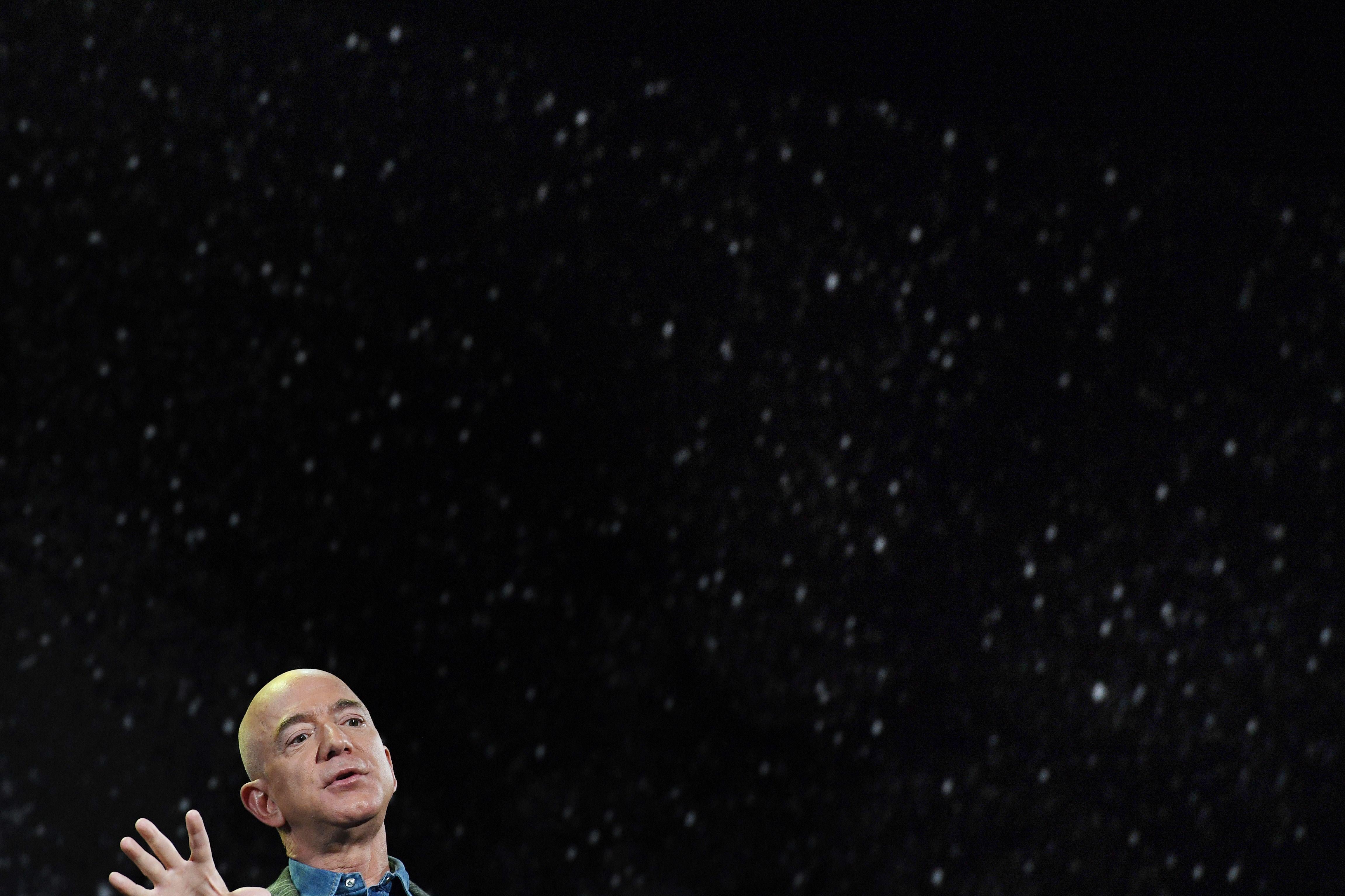 Jeff Bezos raises one hand while speaking, in front of a background of stars.