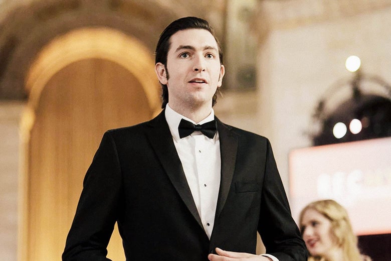 A man in a tuxedo and slicked back hair stands up in a ballroom.