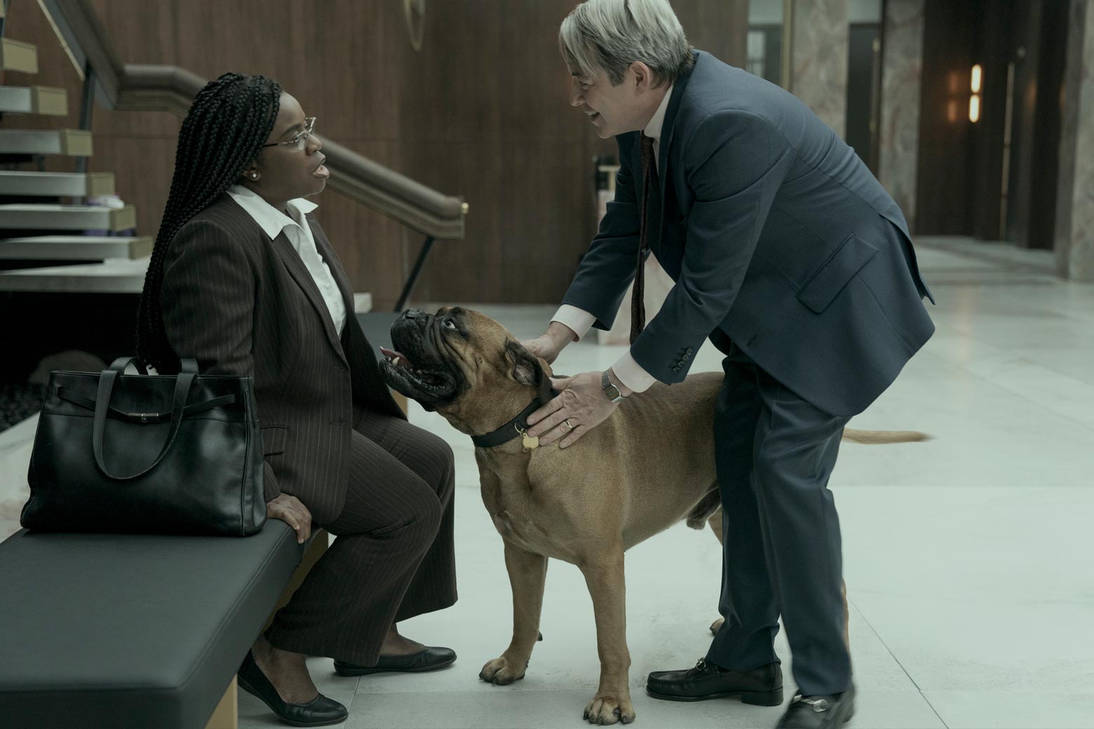 Fictionalized Richard Sackler petting his large mastiff, who has approached Edie Flowers on a bench.