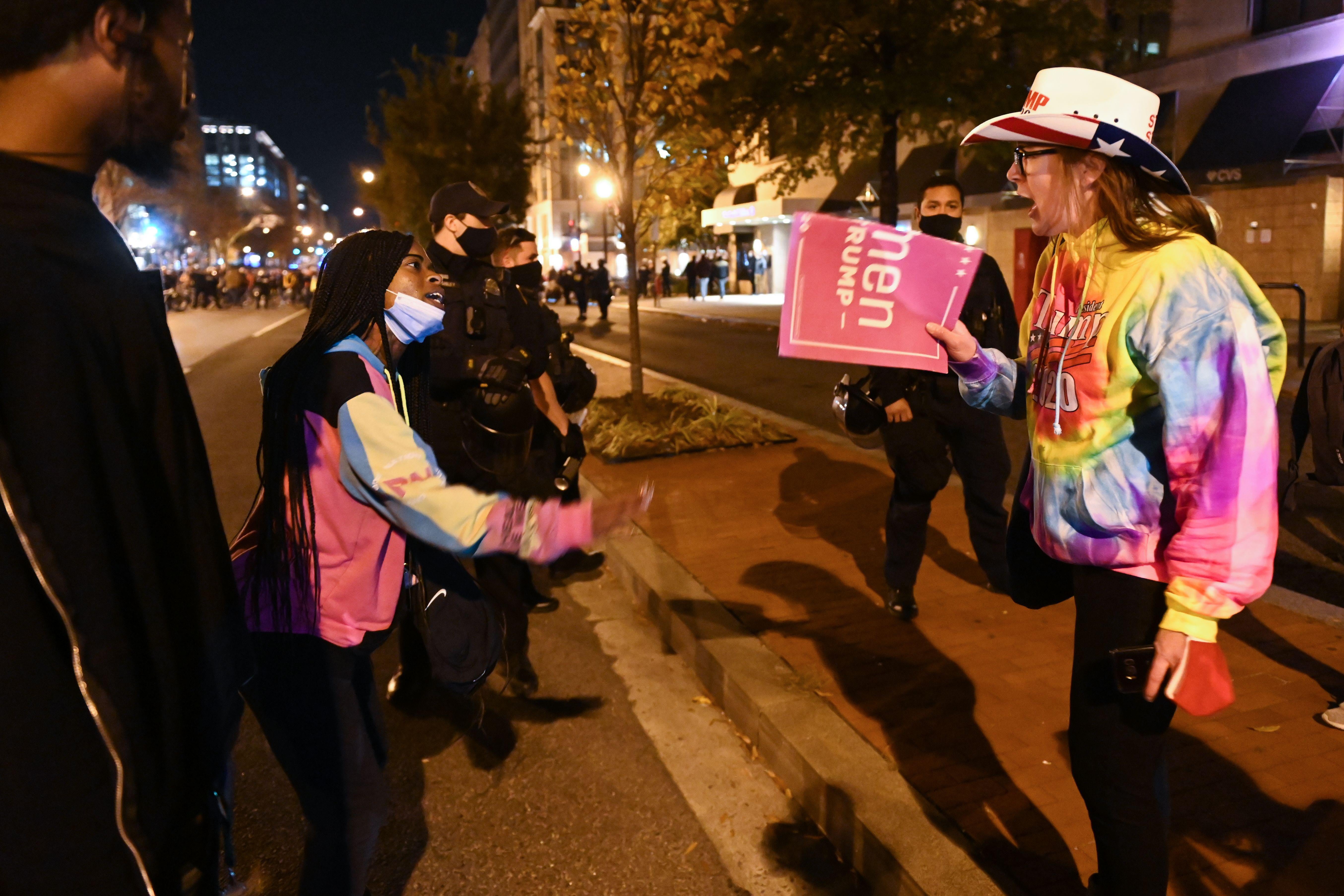 Two people shouting at each other: A Black woman with a mask pulled down to her chin and a white woman with a Women for Trump sign, in American flag cowboy hat and tyedye sweater