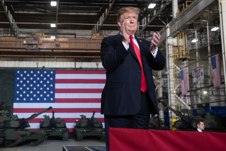 Trump stands in front of a giant American flag and a number of tanks.