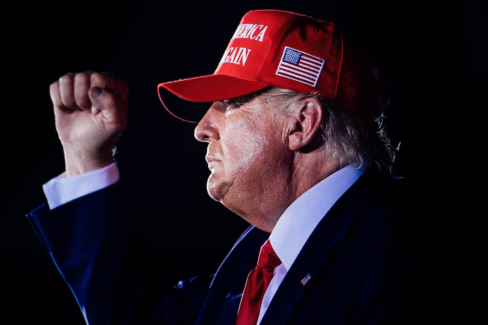 Trump in profile, wearing a red MAGA hat and raising his fist