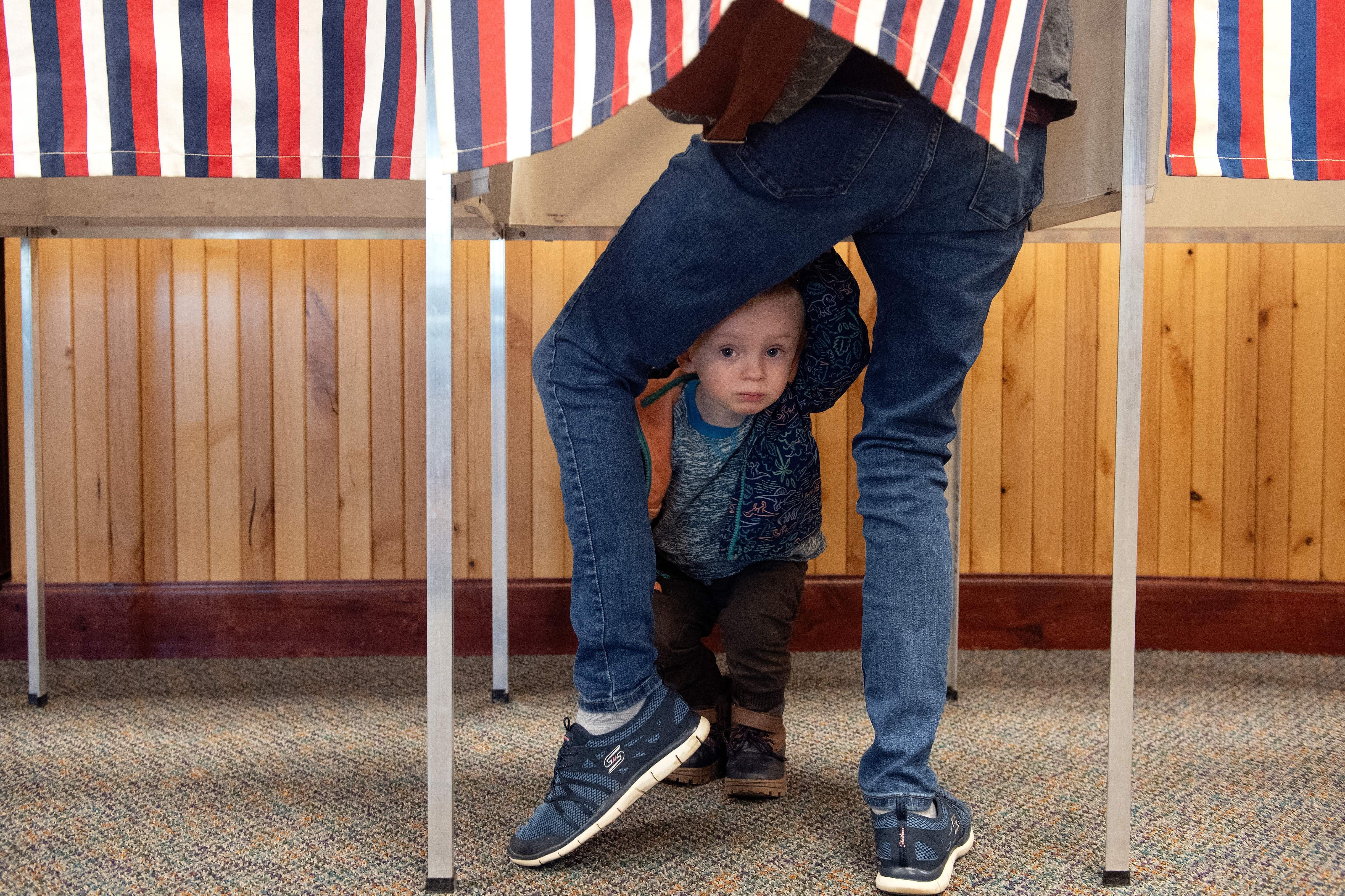 A child peeks out from between his parent's legs as the parent casts a ballot in a voting booth in a wood-paneled room