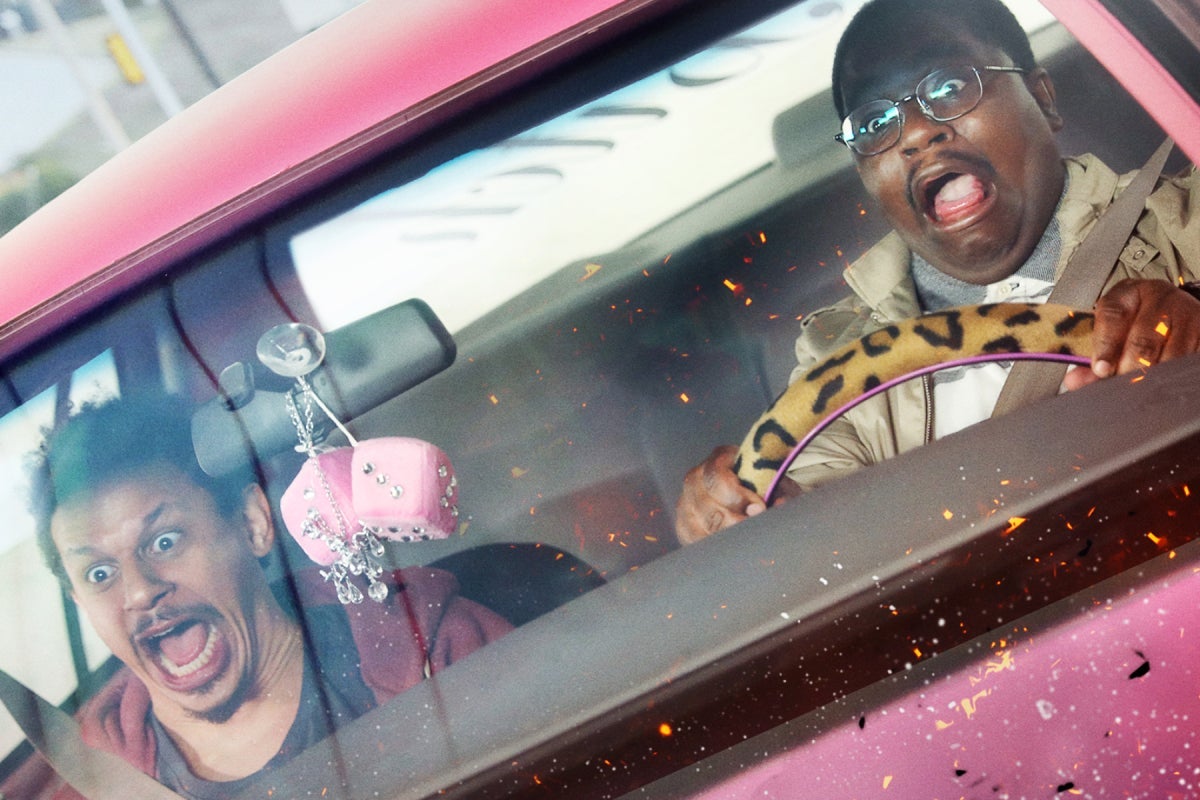 Two men sit in the front of a pink car that has pink fuzzy dice dangling from the rearview mirror and a cheetah print-covered steering wheel. They are both yelling, eyes wide, as flames begin to engulf the front of the car. 