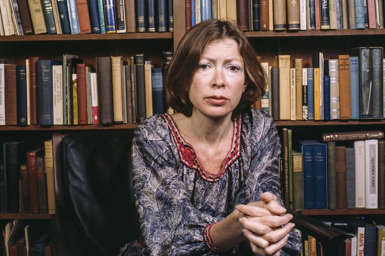 Didion sitting in front of a full bookshelf with her hands clasped in front of her, looking into the camera