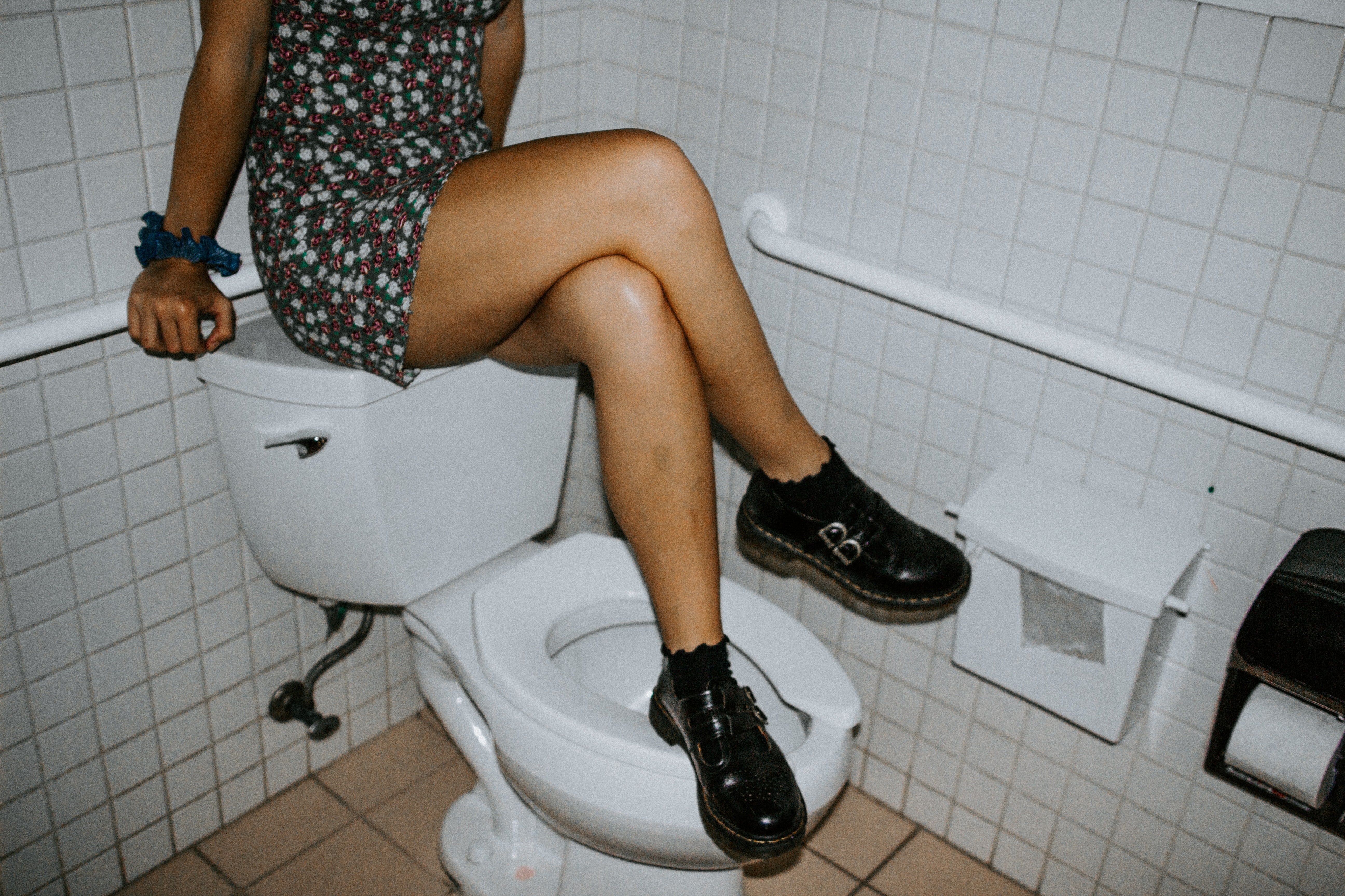 A woman in a dress and boots sitting on the tank of a toilet with her legs crossed. One foot is on the toilet seat. It's a weird place to sit! 