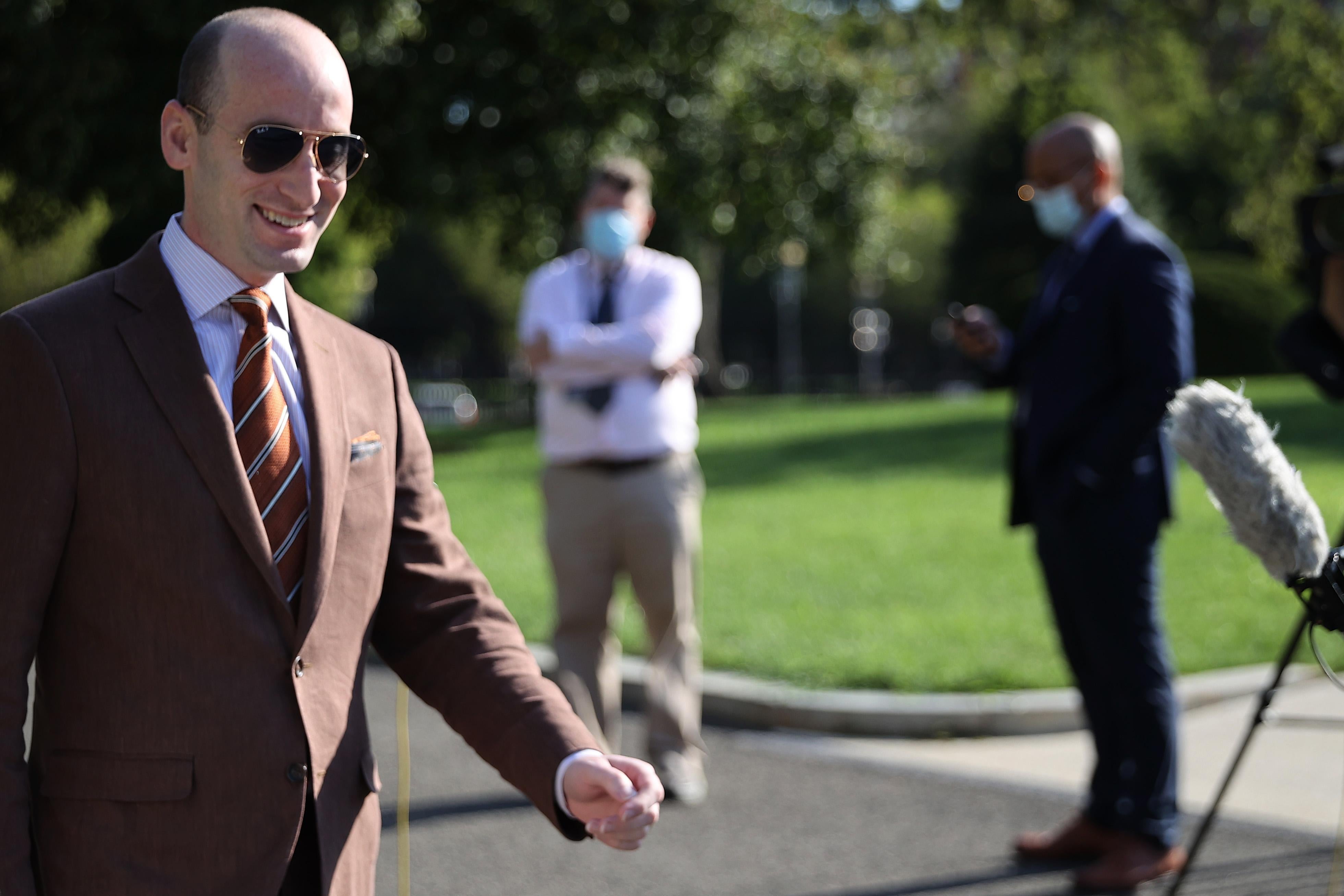 Stephen Miller, wearing a brown suit and aviator sunglasses, smiling widely as he walks