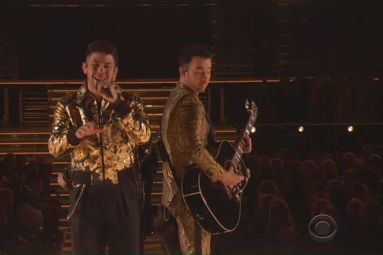 Nick Jonas sings into a microphone at the Grammys as Kevin Jonas stands beside him, playing guitar.
