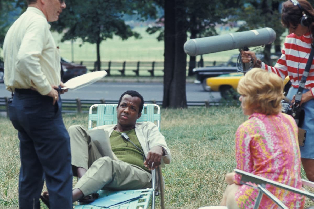 A Black man sits in a lawn chair with two white actors and a white sound man standing around him.