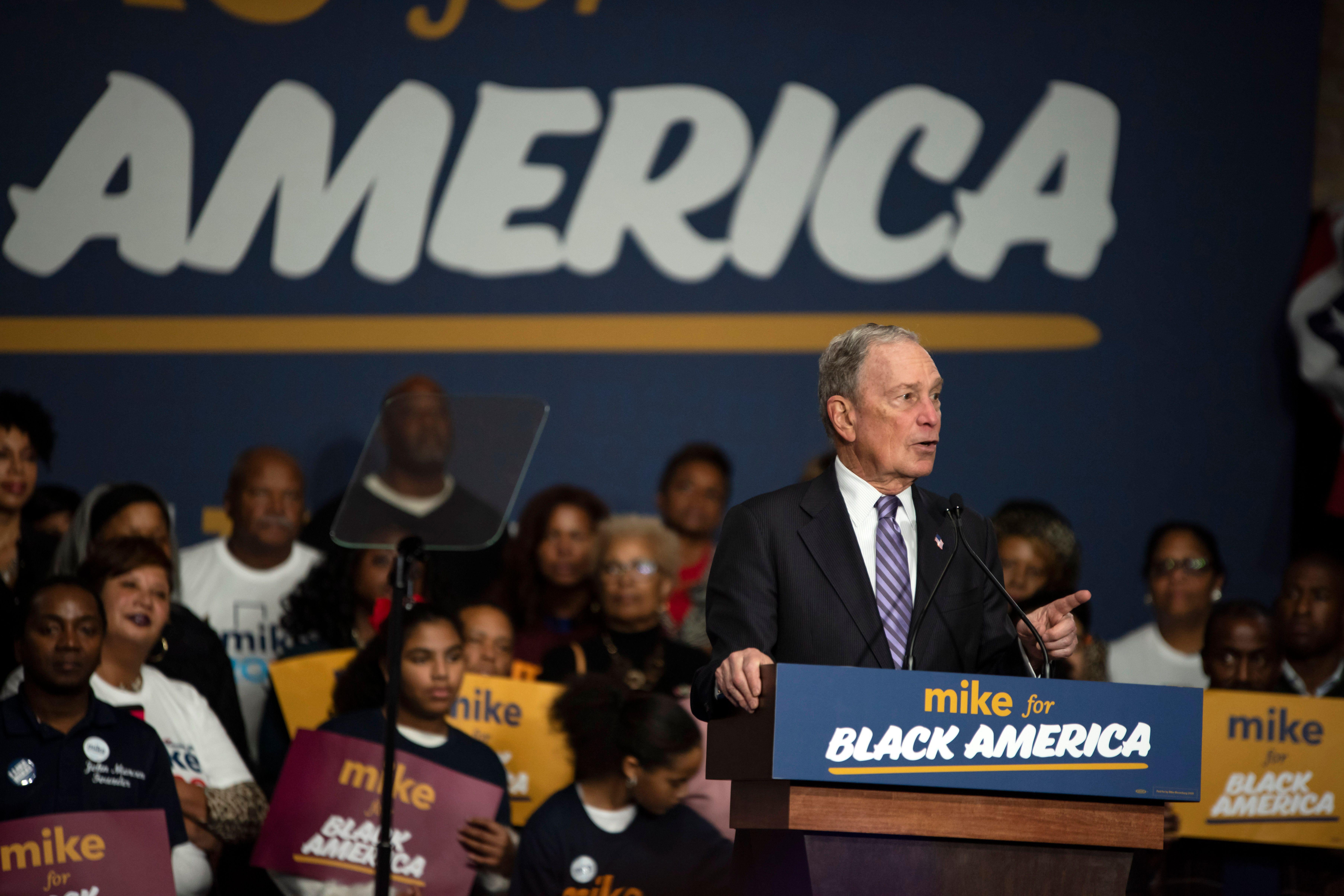 Michael Bloomberg speaks into a podium that says "Mike for Black America," as black supporters sit behind him.