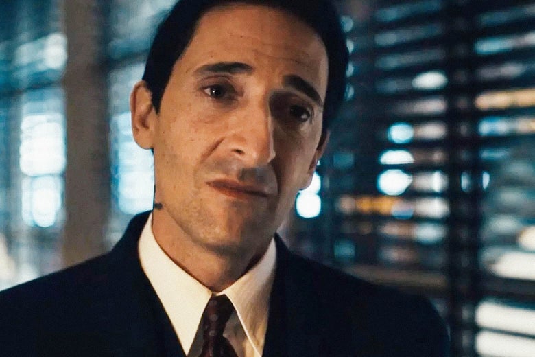 Close-up on a man in a suit titling his head to the side in front of a window with blinds on it.