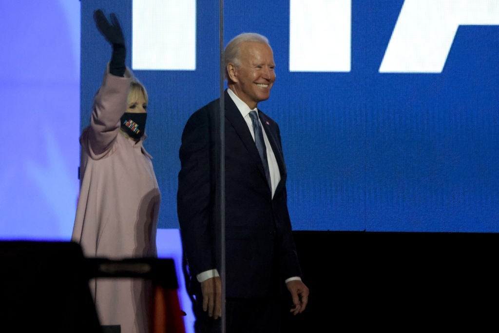 Biden smiles in front of a large blue screen as his wife Jill, standing beside him, waves.
