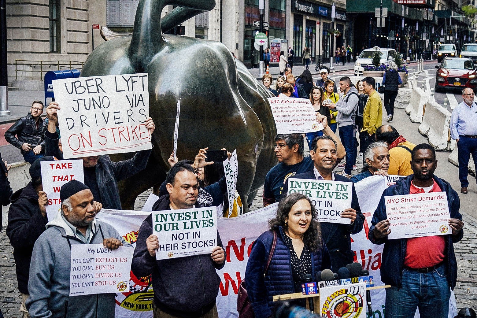Next to the Charging Bull, protesters hold signs with slogans such as "INVEST IN OUR LIVES NOT IN THEIR STOCKS," "UBER LYFT JUNO VIA DRIVERS ON STRIKE," and "YOU CAN'T EXPLOIT US."