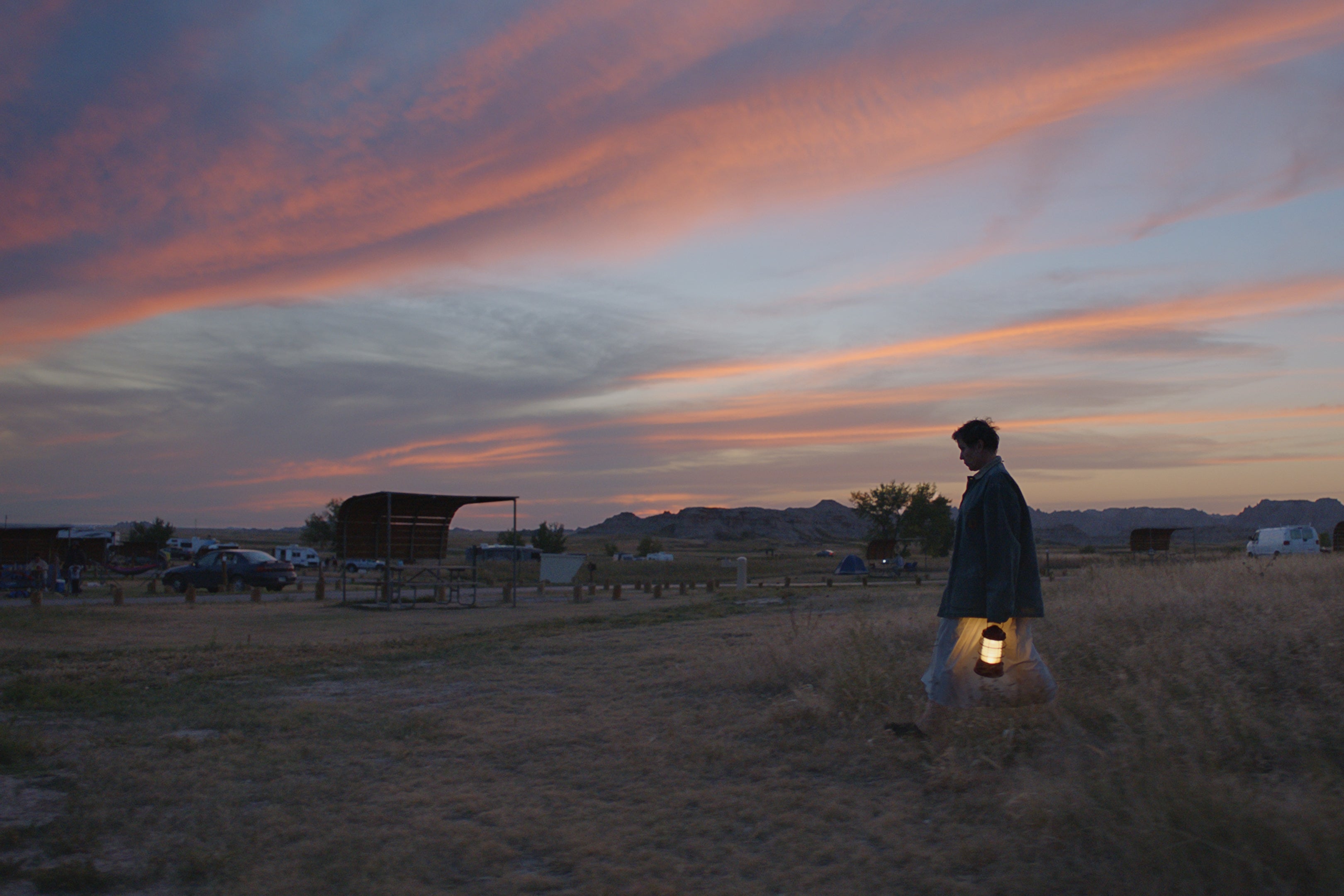 She walks through the brush near a rural parking lot against a colorful sky, carrying a lantern.
