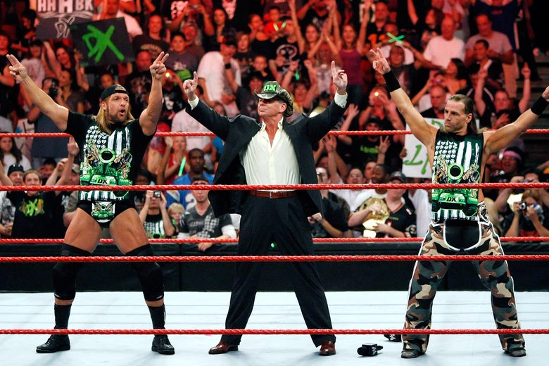 Two wrestlers stand on either side of Vince McMahon arms raised in the middle of the ring during a WWE event.