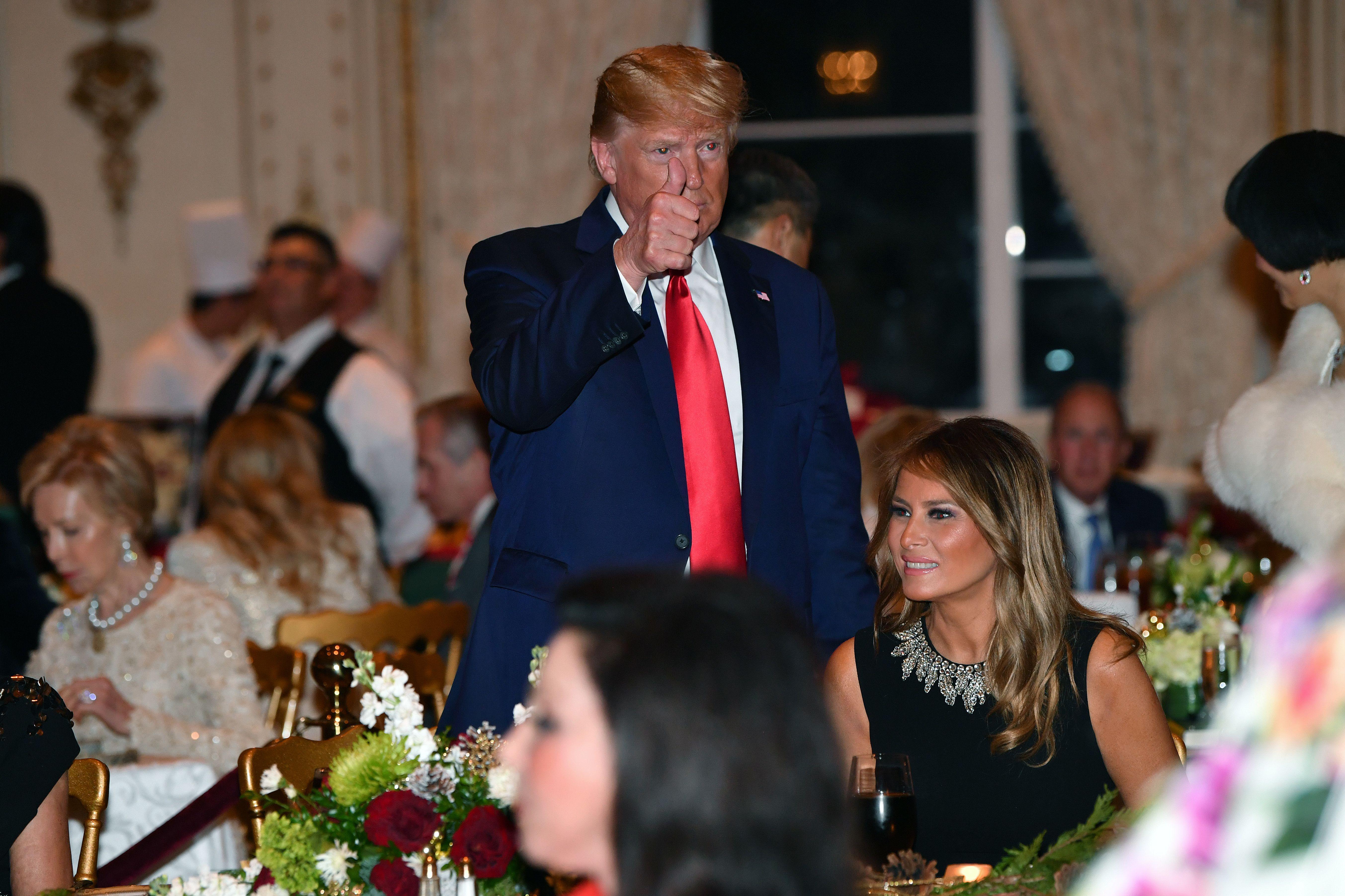 Trump, standing, makes a thumbs-up. Melania is seated next to him in a fancy banquet hall full of other guests.