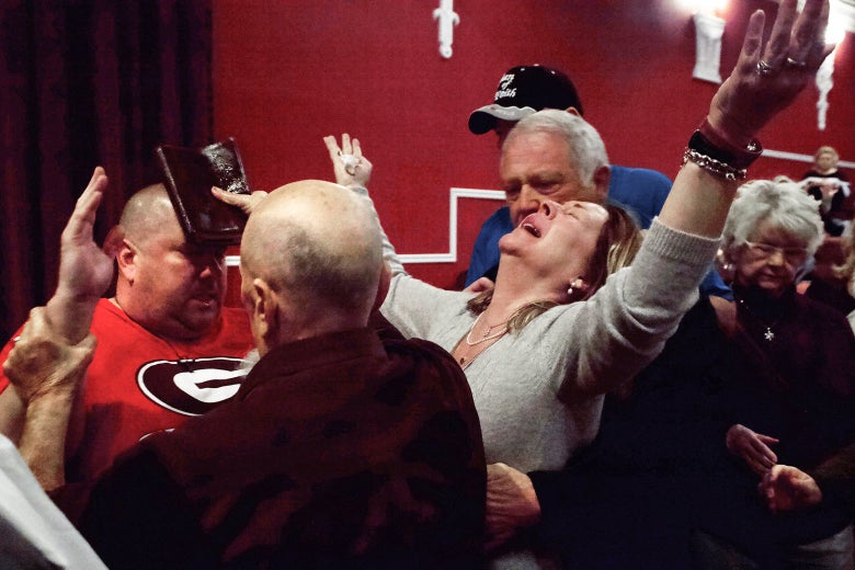 A woman raises her hands, one carrying an oily Bible, as she collapses back into a man's arms, with others surrounding her.