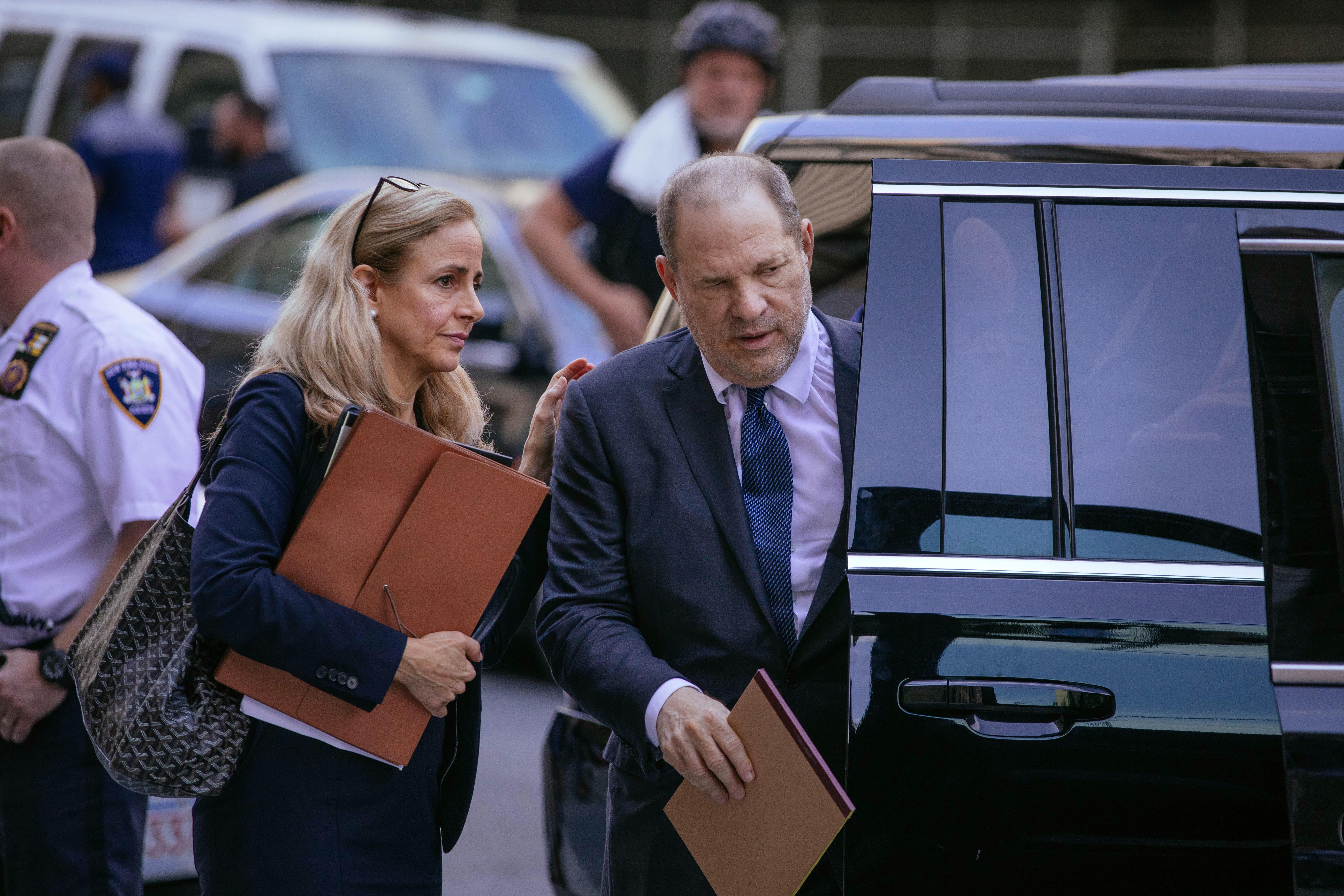 Harvey Weinstein entering a large black vehicle. A woman holding a folder touches his shoulder.