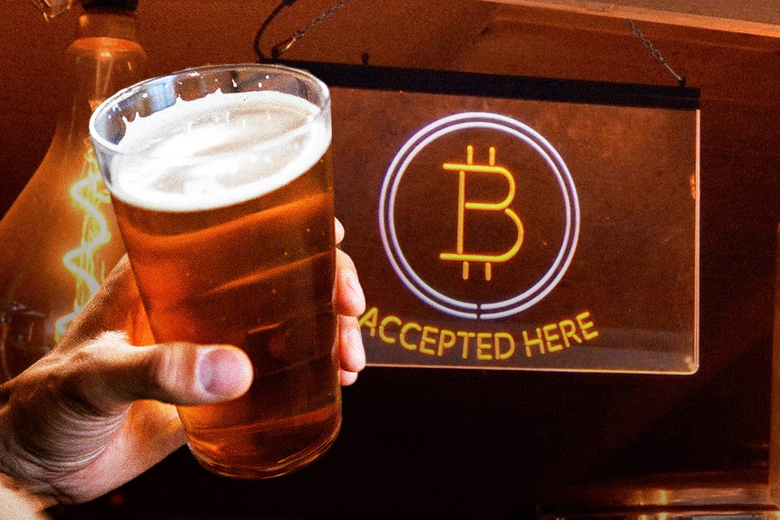 A white hand holds a beer in front of a sign with the Bitcoin logo that says "Bitcoin accepted here."