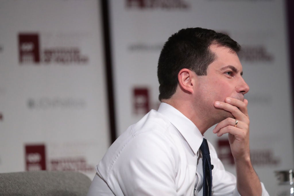 Buttigieg, wearing a white shirt, holds his chin pensively while seated on a stage.