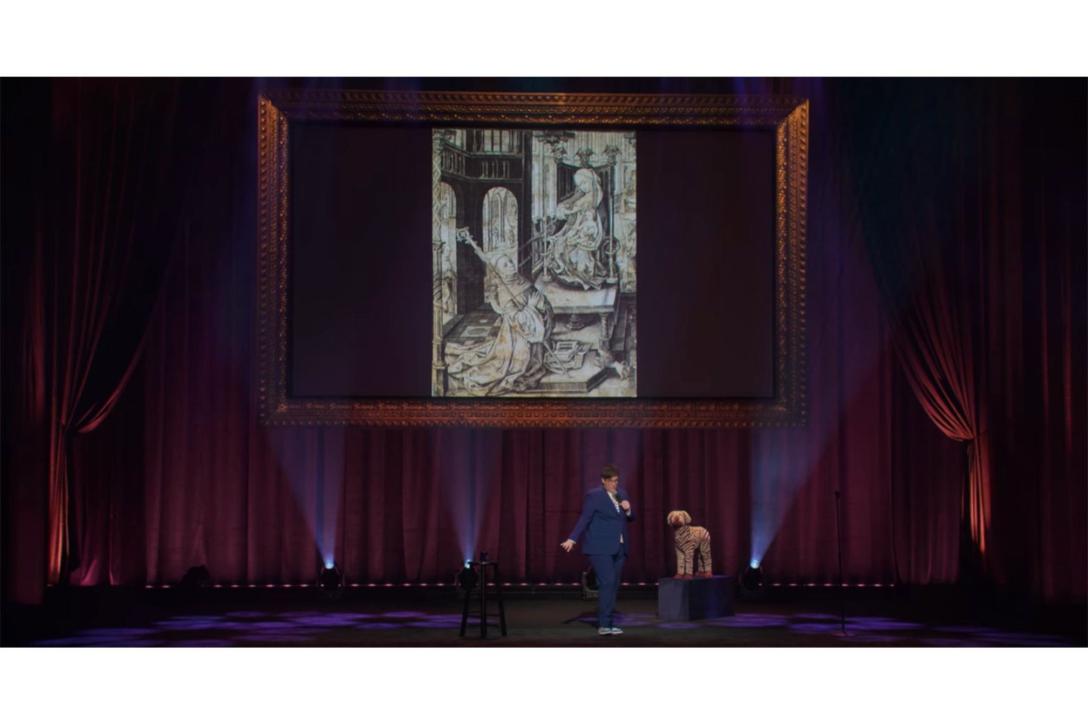 Hannah Gadsby stands onstage in front of a projection of an engraving of St. Bernard kneeling while Mary lactates into his mouth.