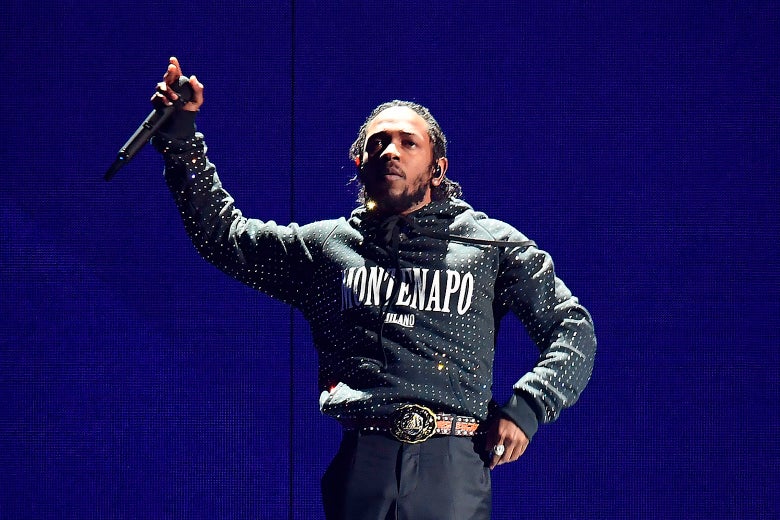 Kendrick Lamar onstage wearing a shiny black hoodie holding a mic up in his right hand