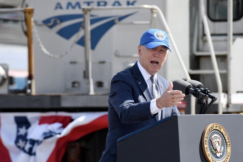 Joe Biden wears a blue hat bearing the presidential seal and gestures as he speaks at a podium at the 30th Street Station in Philadelphia, with the Amtrak logo and American flag bunting behind him
