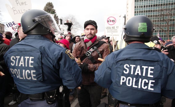 Michigan State Police in riot gear stand with batons while protestors block a street during a rally at the Michigan State Capitol to protest a vote on Right-to-Work.