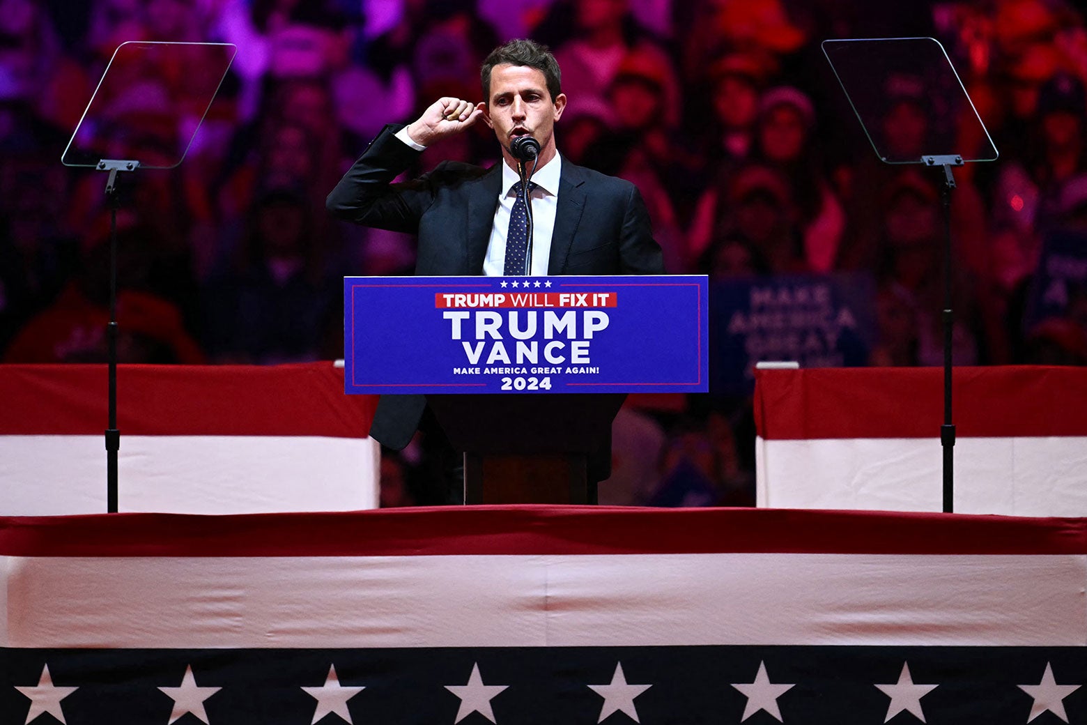 Tony Hinchcliffe in a conservative suit speaking from behind a lectern with Trump Vance 2024 on it.