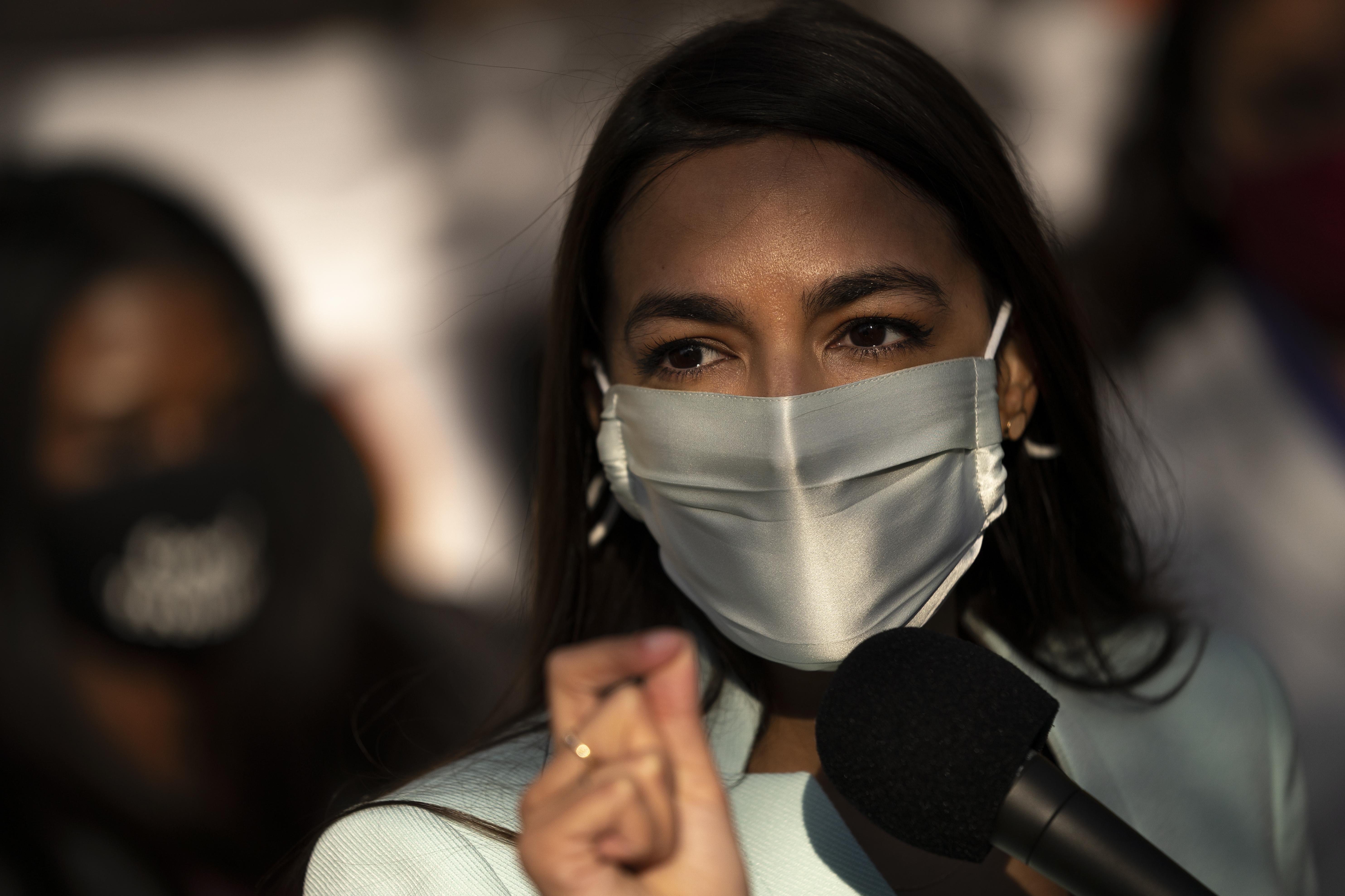 Alexandria Ocasio-Cortez speaking with a mask on at a mic outside
