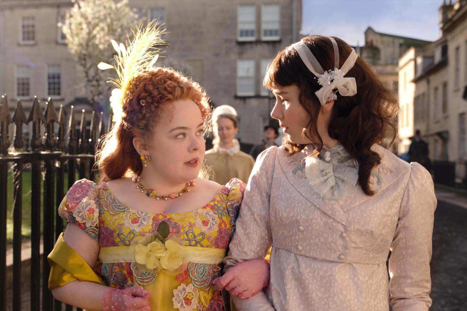 Two women in period dress link arms as they walk outside.
