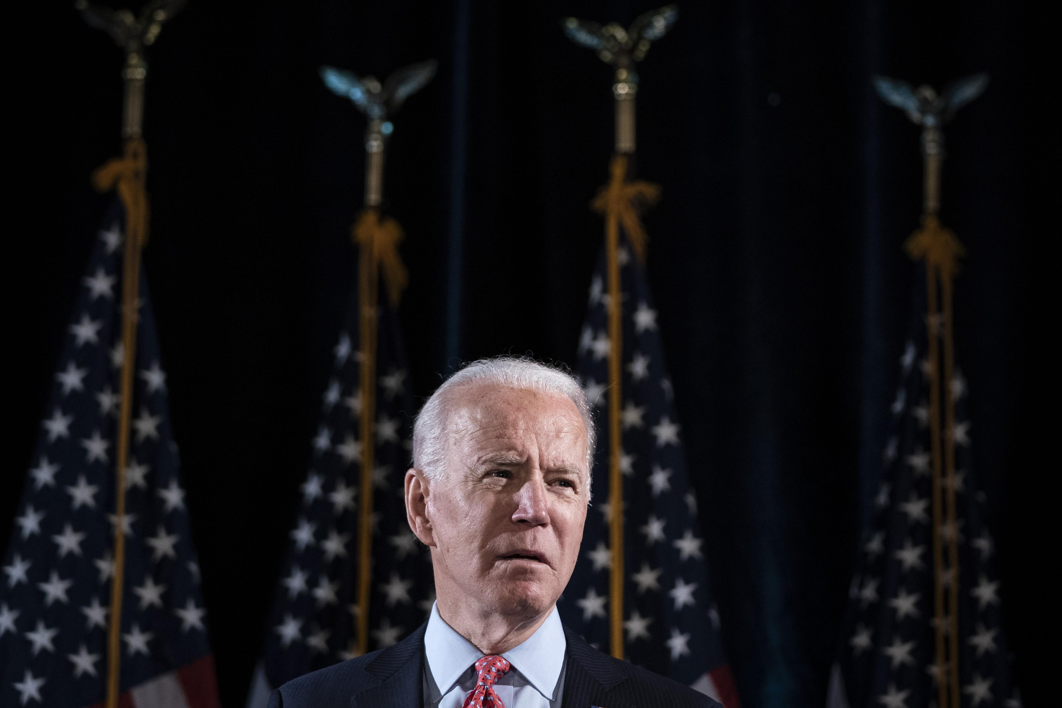 Joe Biden stands in front of four American flags. 