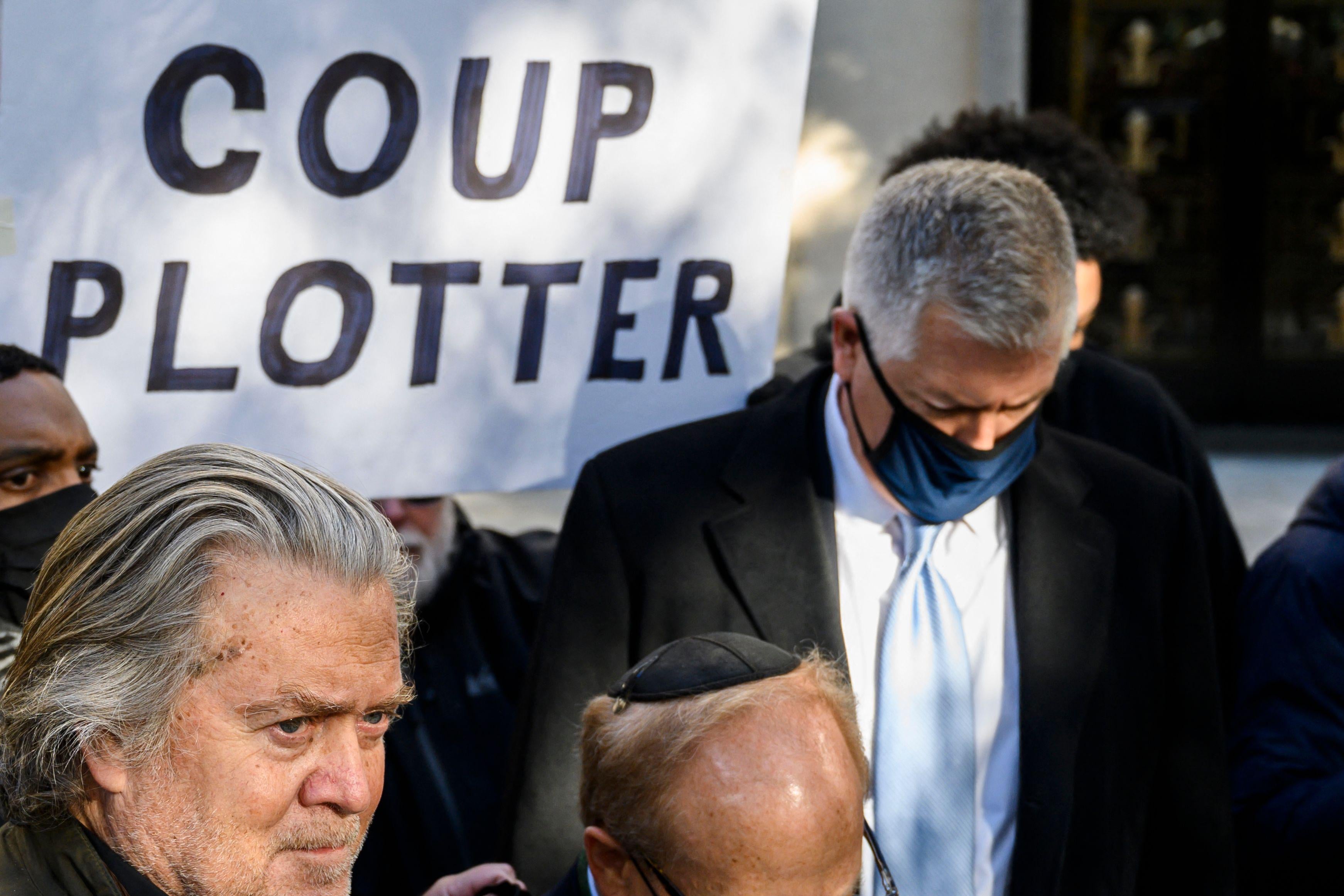 Bannon stands outside among other people while behind him someone holds up a sign that says "COUP PLOTTER"