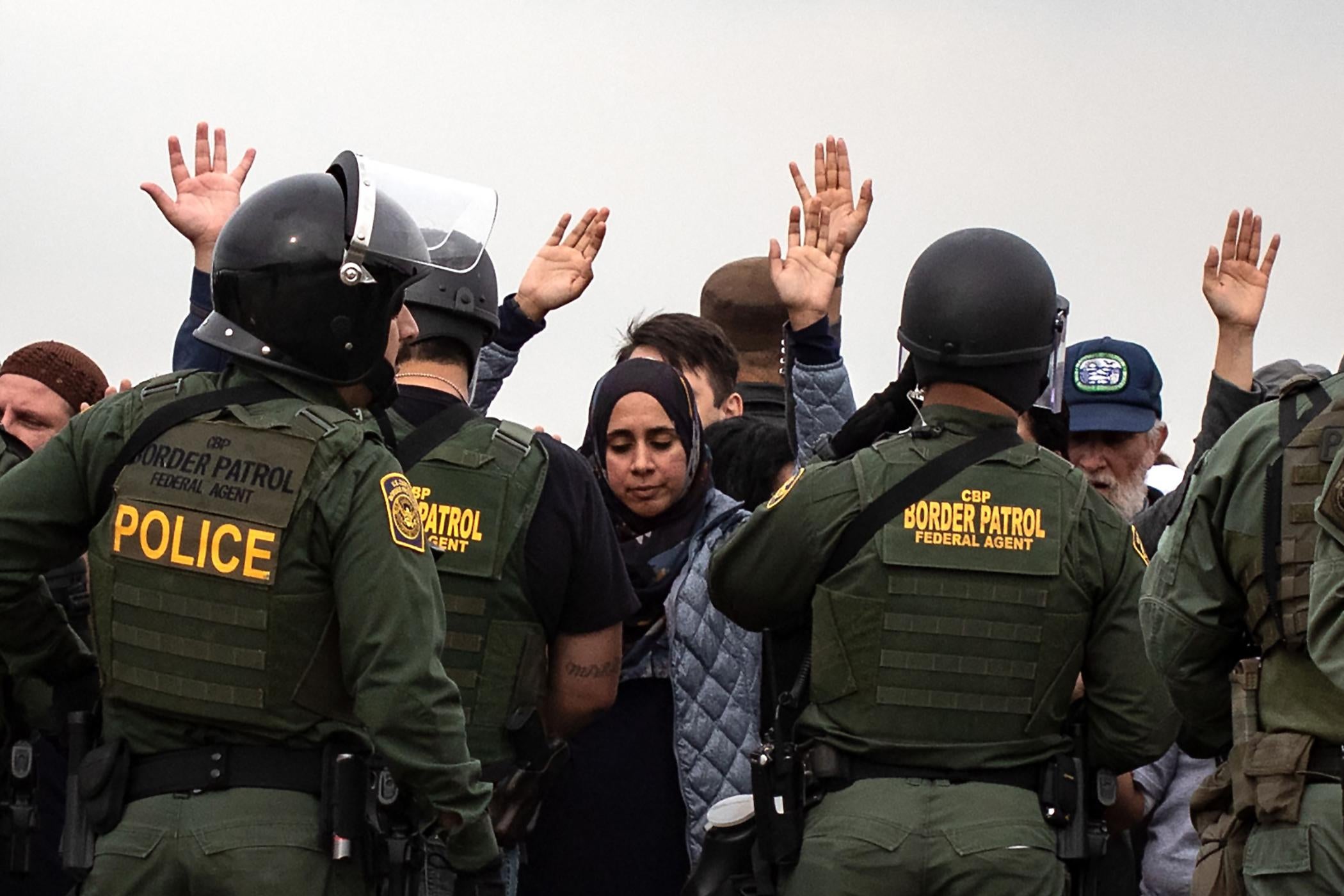 Demonstrators look down and hold their hands in the air. A line of border patrol agents in uniform stand in front of them.
