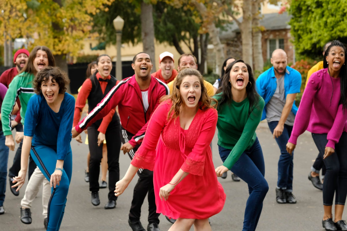 Rachel Bloom leads a crowd of people dancing in a street, all wearing bright colors.