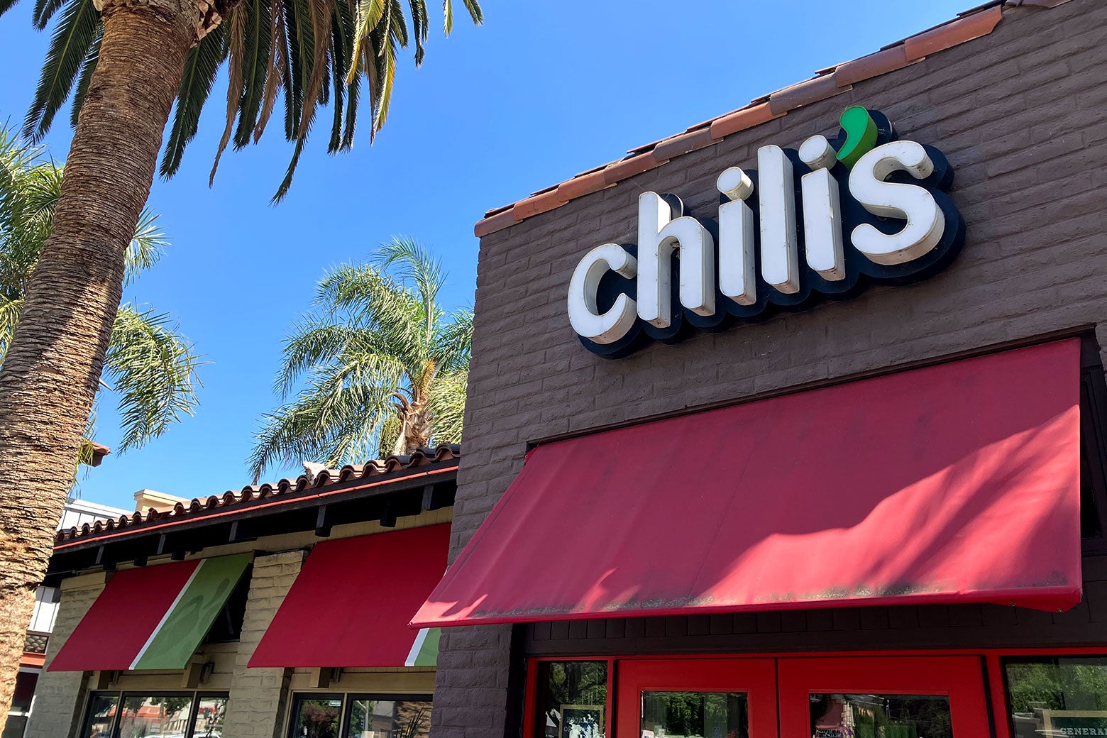 Exterior of a Chili's featuring a red awning, palm trees, and the company's iconic chile pepper logo.