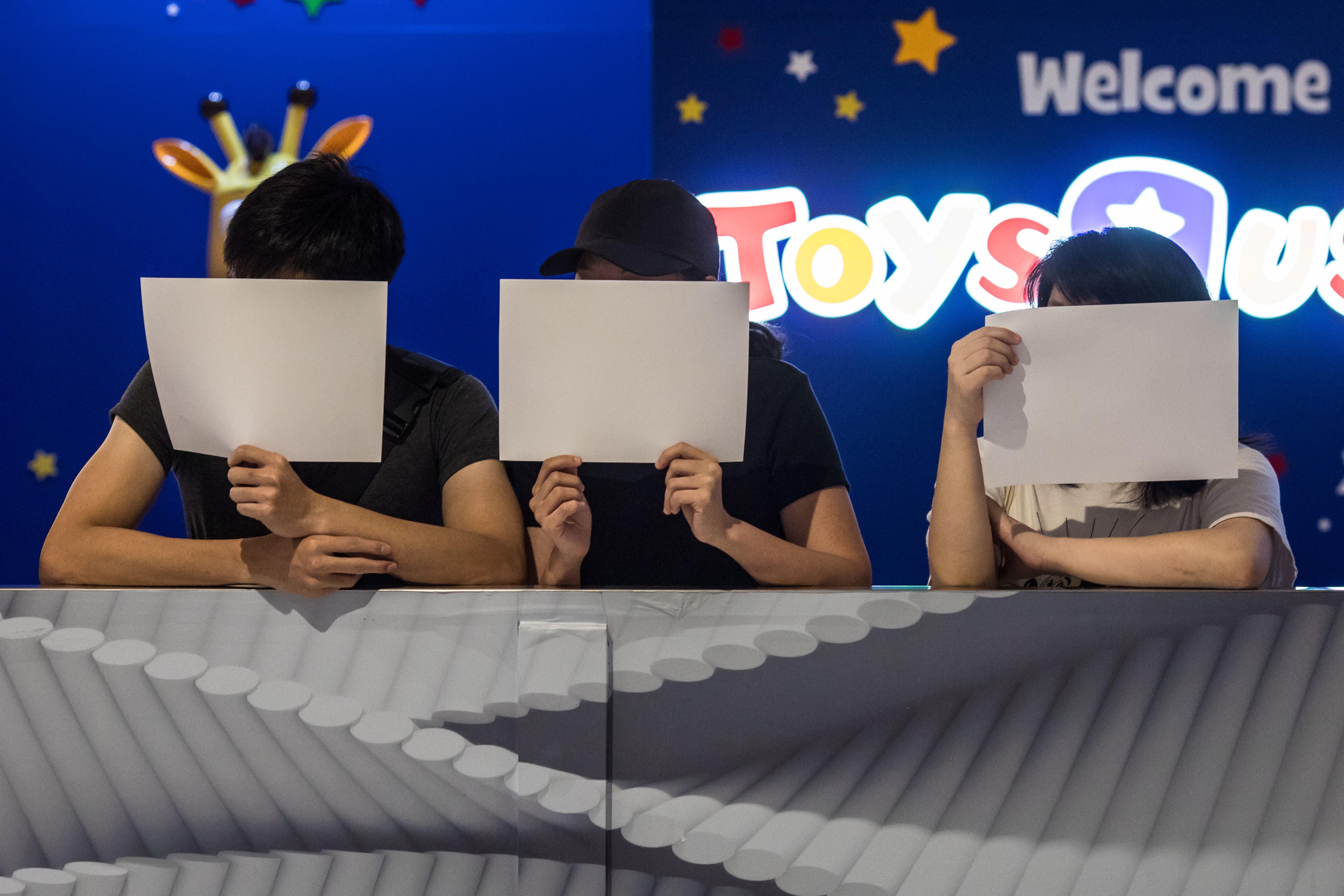 Three protesters hold up white pieces of paper in front of their faces while standing in front of a Toys 'R' Us.