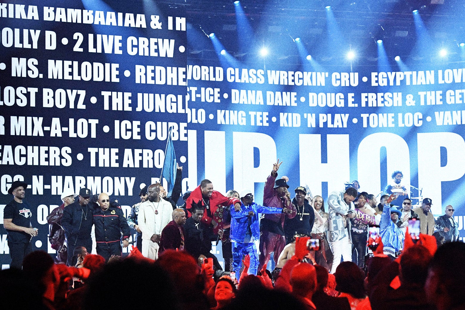 A group of 15 to 20 rappers perform onstage, with the names of hip-hop legends in the background.