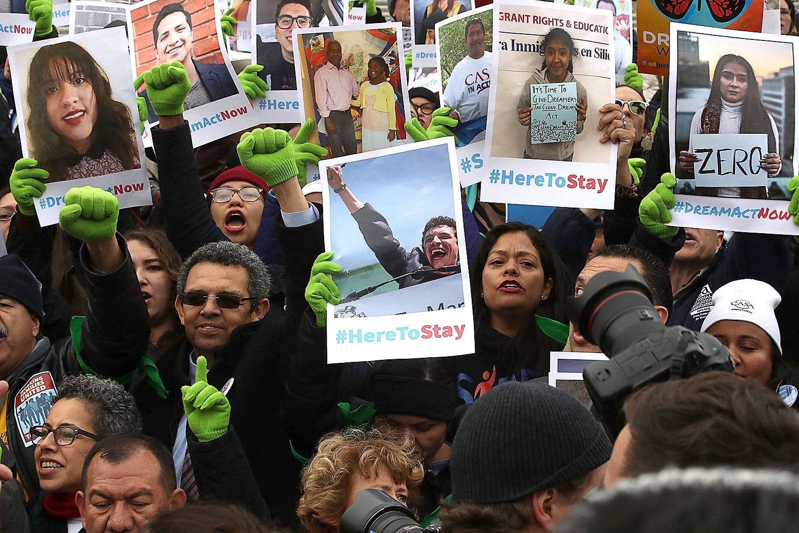 A crowd of protesters holding up posters with photos of Dreamers and the hashtag #HereToStay.