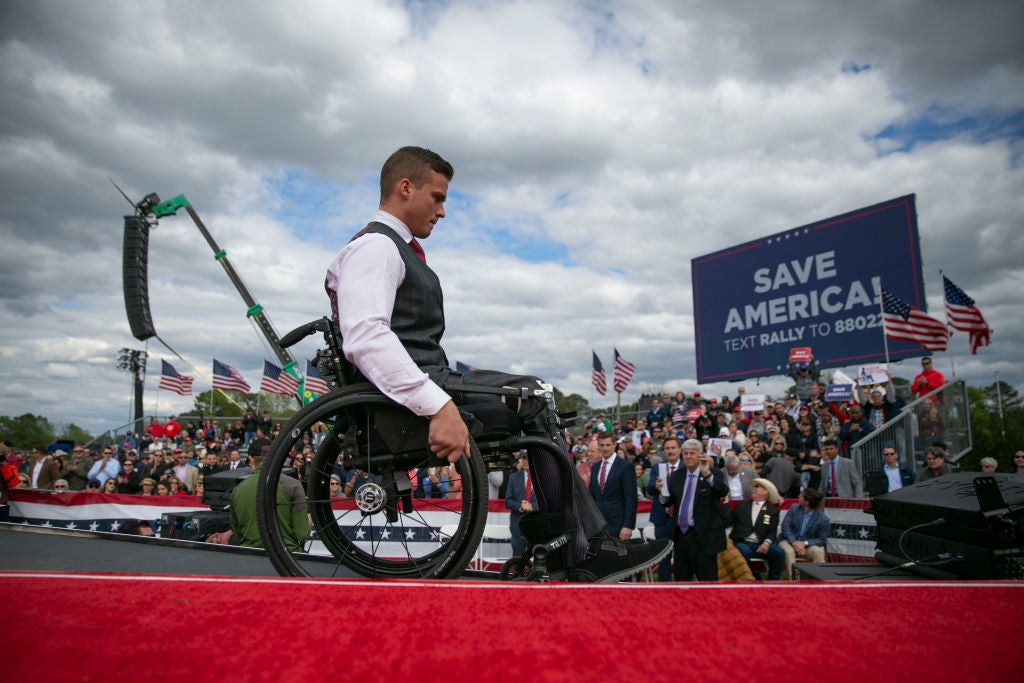 Cawthorn, seated in a wheelchair and wearing a gray vest with a red tie, moves toward the right of the frame down a ramp off a red carpeted stage. A rally crowd and a "Save America" sign can be seen behind him.
