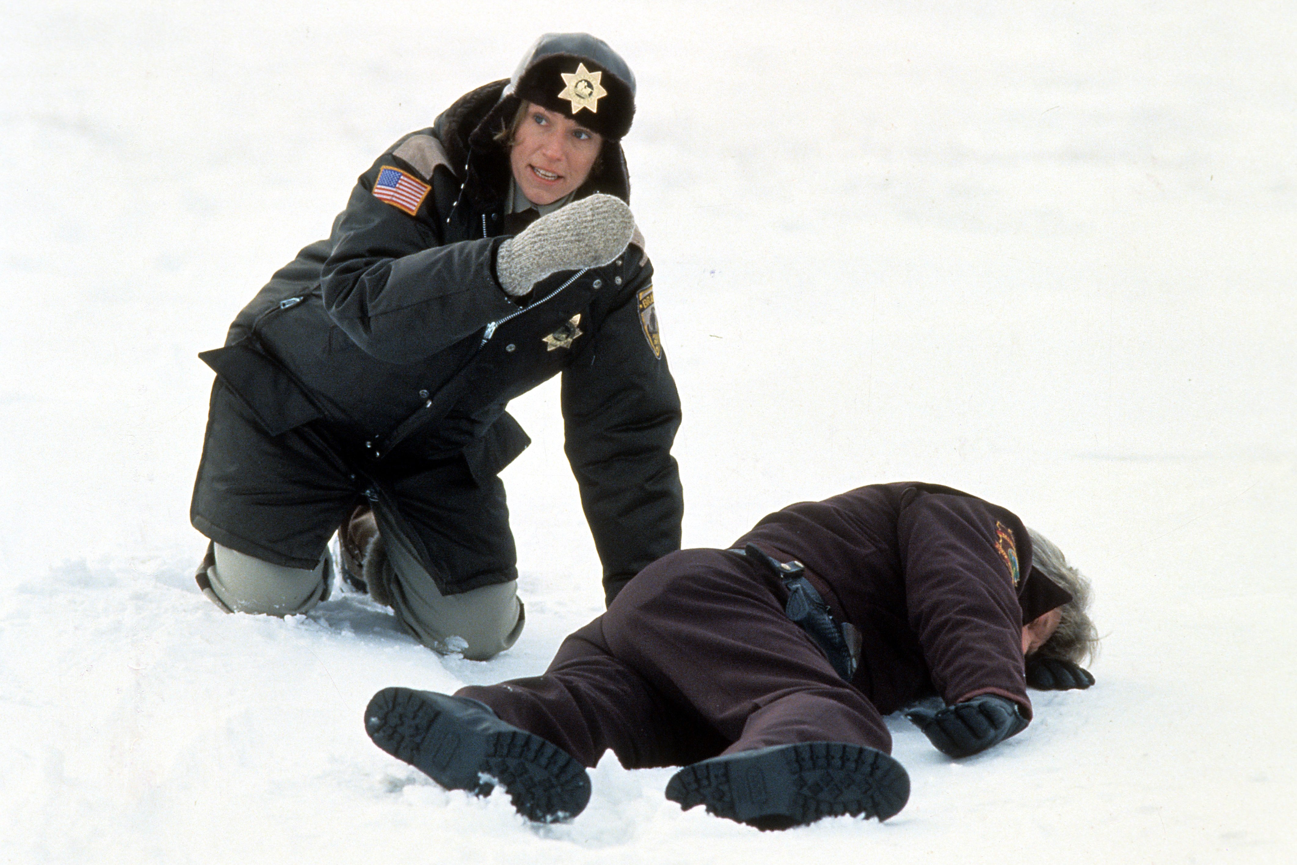 In a snowy field, a female police officer, wearing a winter uniform, kneels over the body of another police officer lying on the ground while looking off screen to something or someone else. 