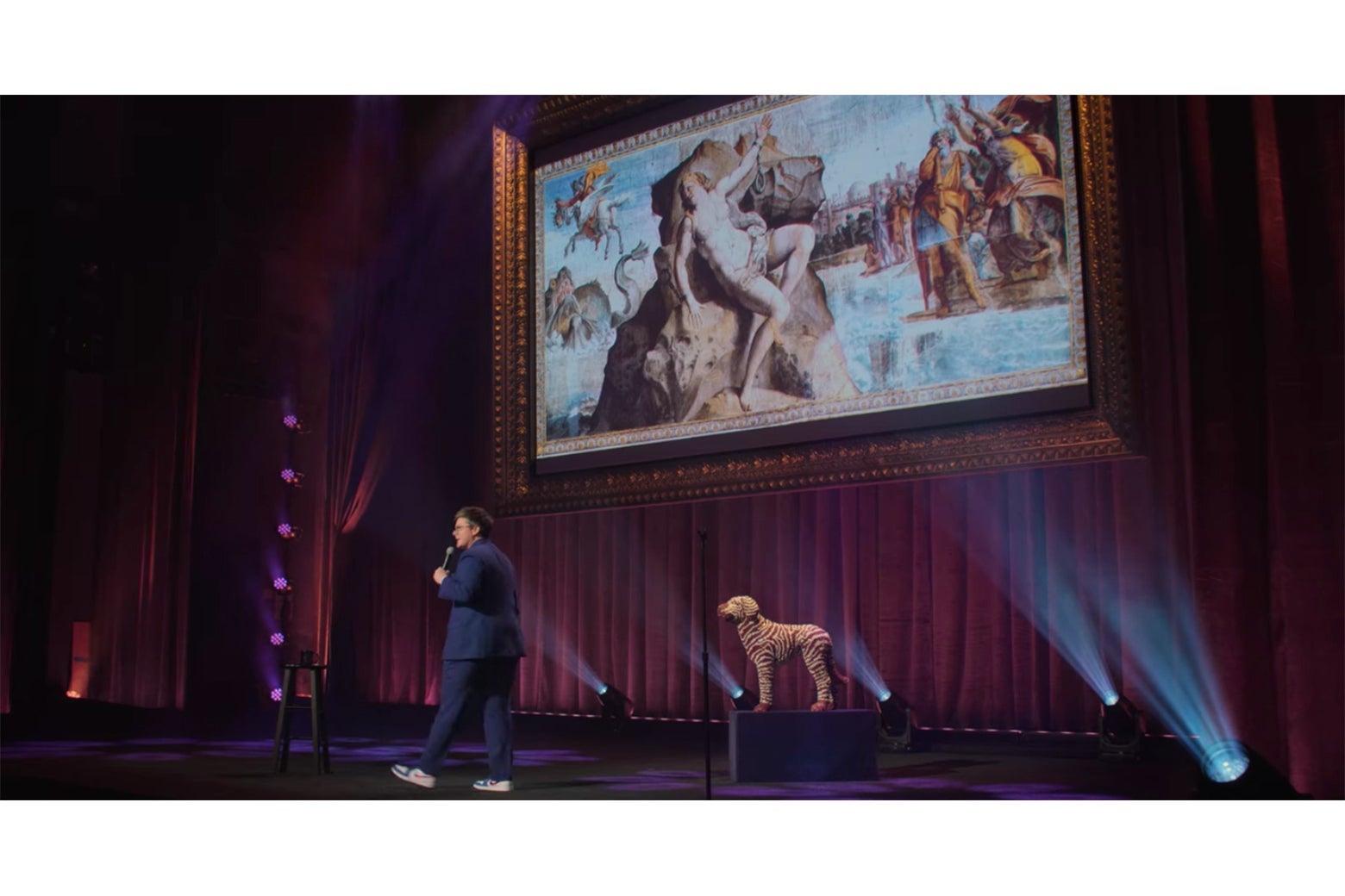 Hannah Gadsby stands in front of a projection screen displaying a fresco of Andromeda chained to her rock.