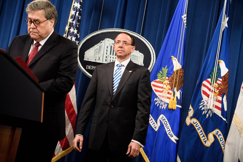 Rosenstein, standing straight with his arms to the side, looks straight forward as Barr speaks at a lectern.