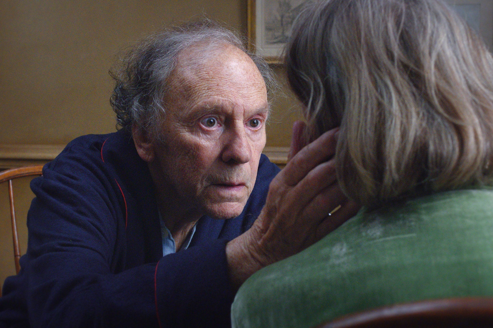 An elderly man, played by Jean-Louis Trintignant, sports wide eyes as he cups the face of an elderly woman whose back is to the camera.