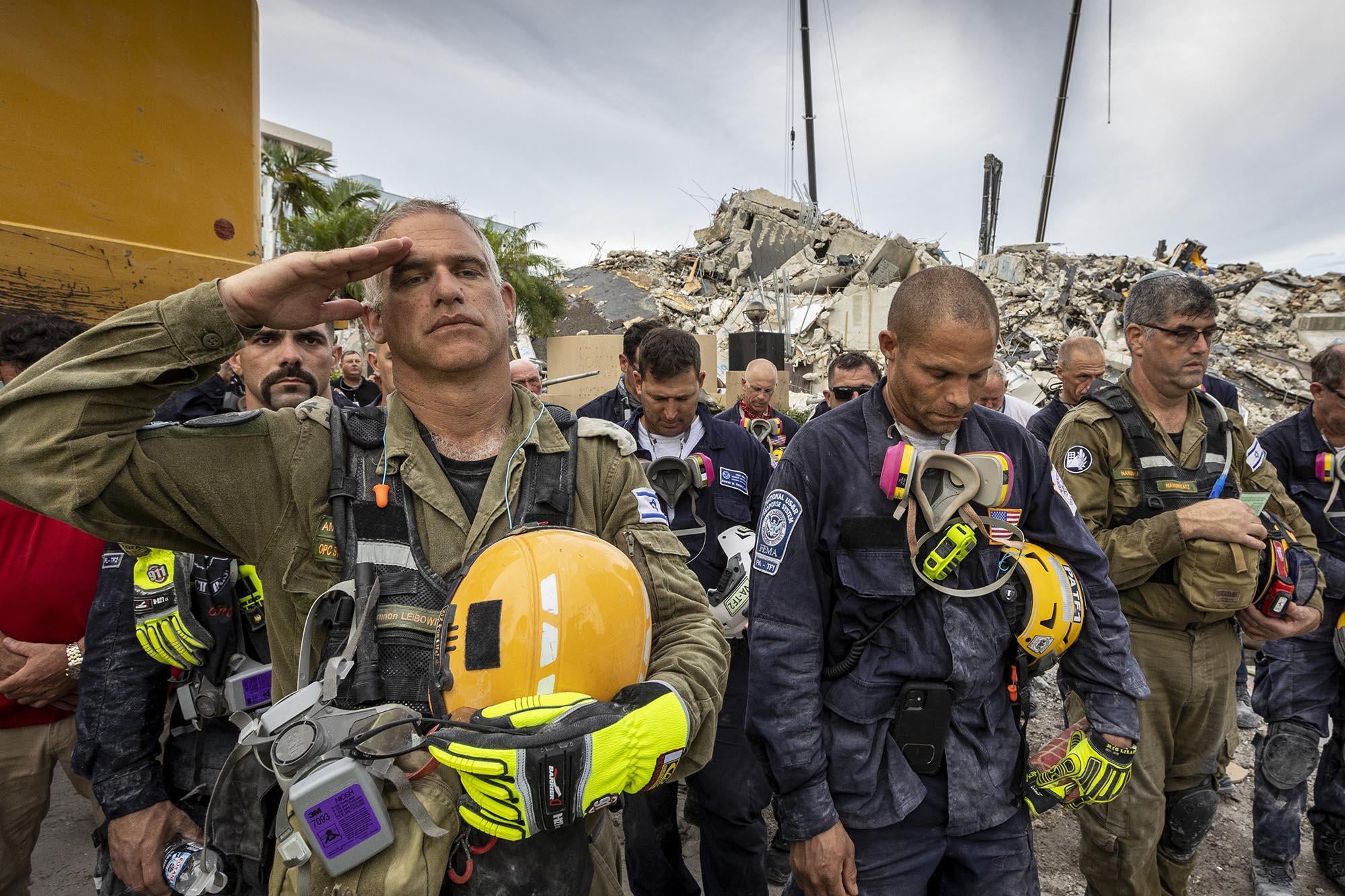 One worker salutes, as members of search and rescue teams gather with rubble in the background for a moment of silence and prayer for the victims of the condo collapse. 