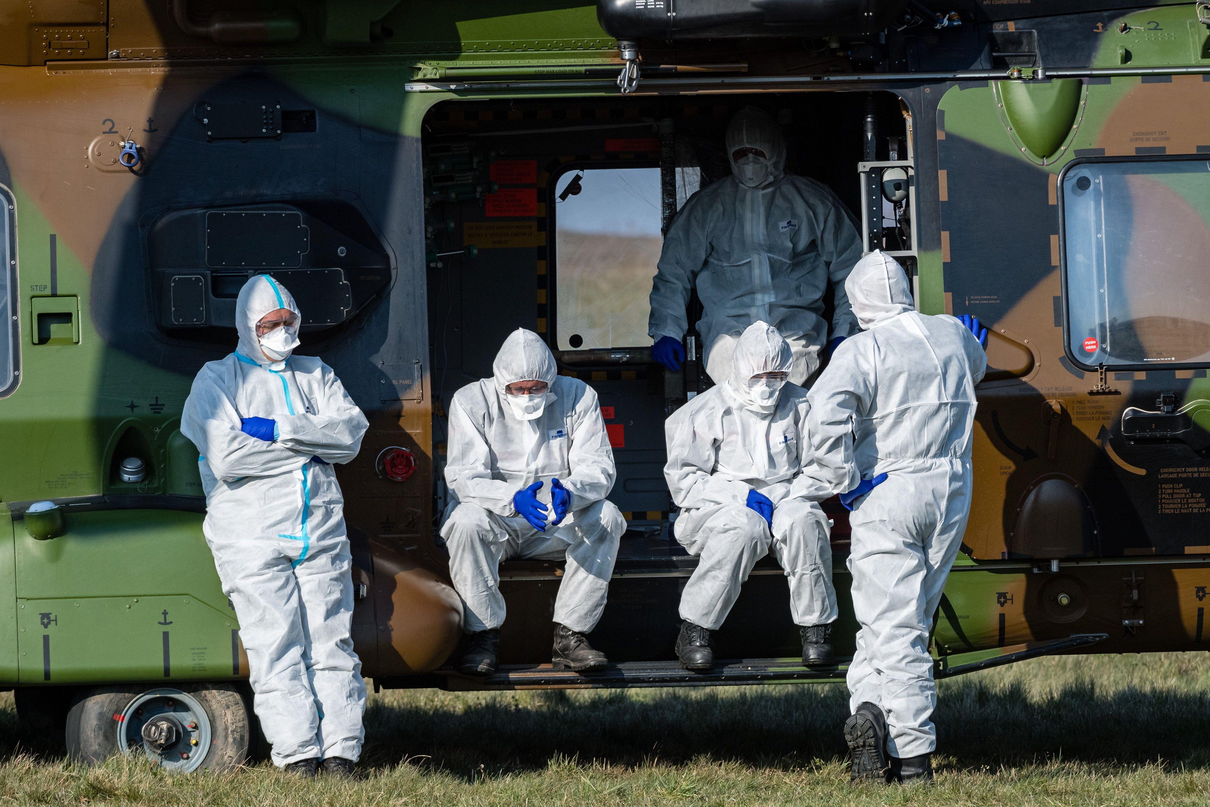 Five people in masks and protective suits are seen inside and in front of a vehicle.