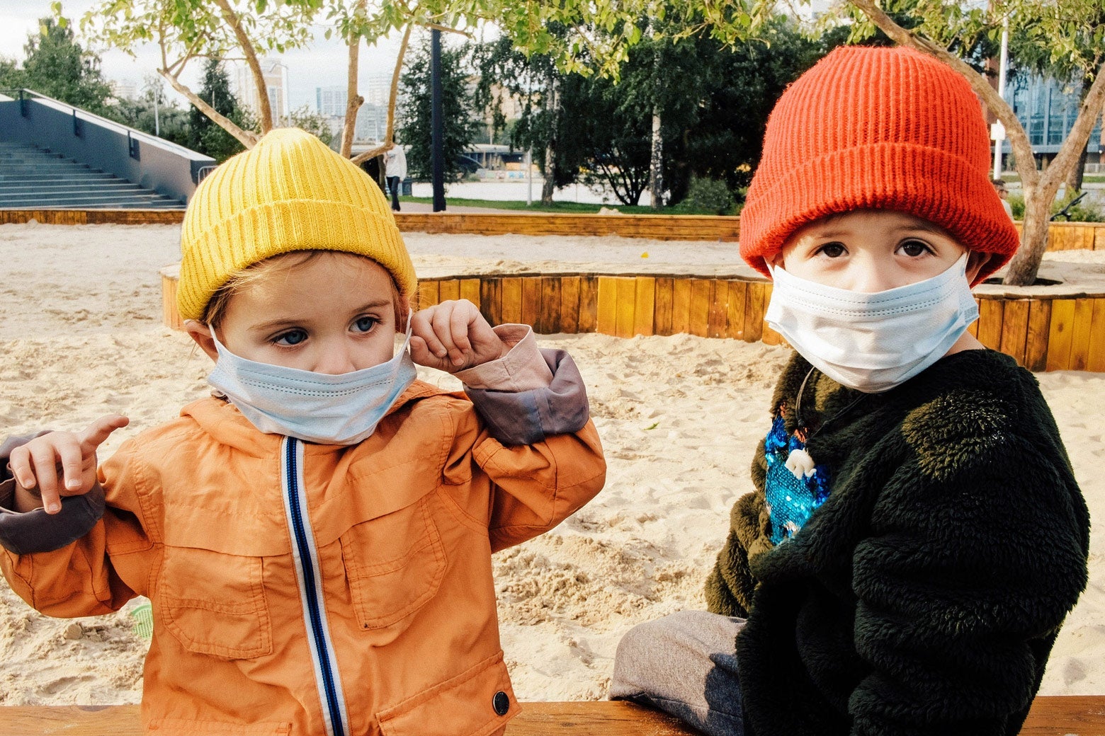 Two little kids in a sandbox wearing masks.