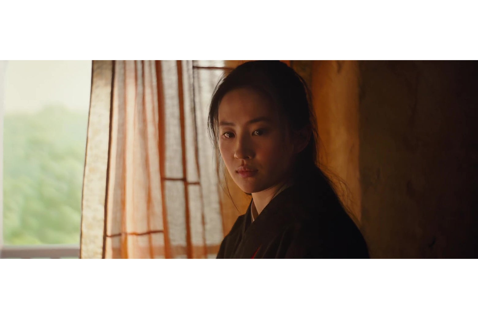 Liu Yifei in a still from Mulan; an overly-bright window and a very dark interior wall are behind her.