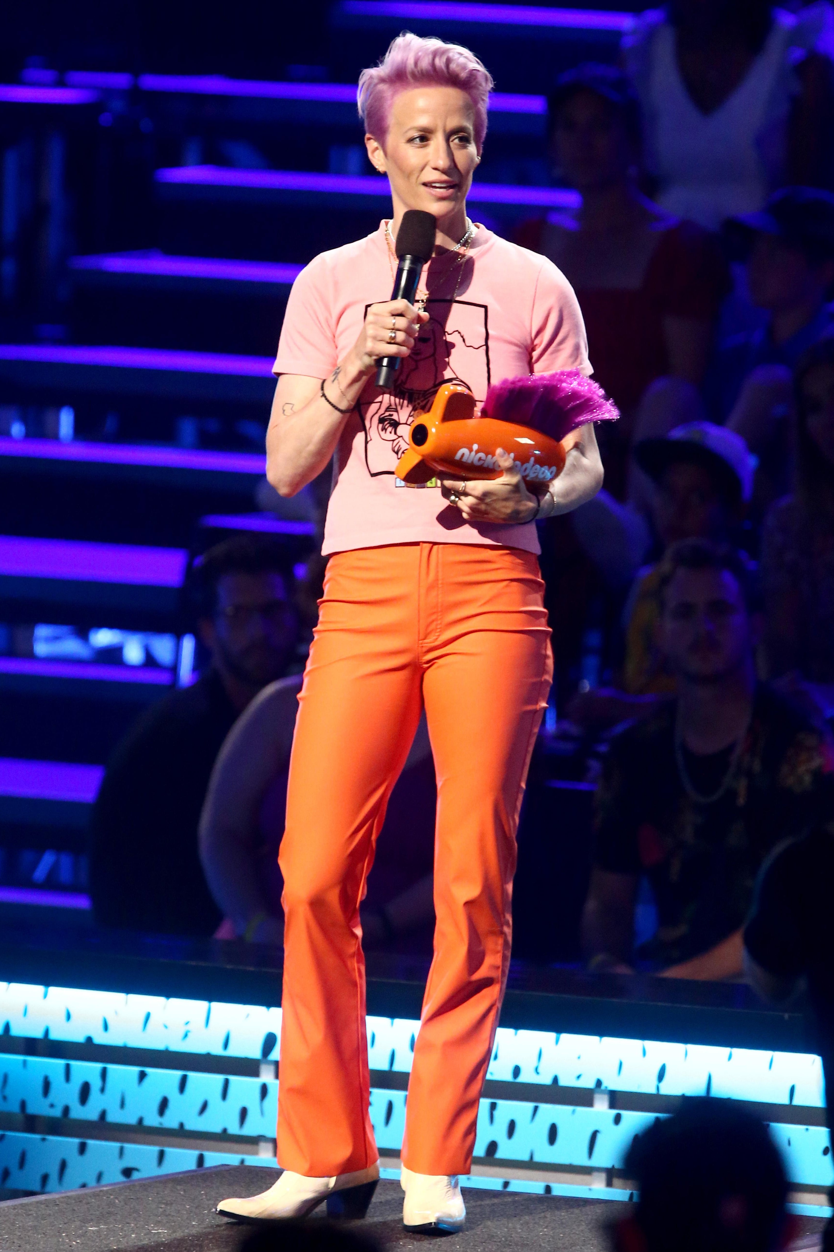 Megan Rapinoe holds an orange Nickelodeon trophy and speaks into a microphone onstage, wearing orange pants and white boots.